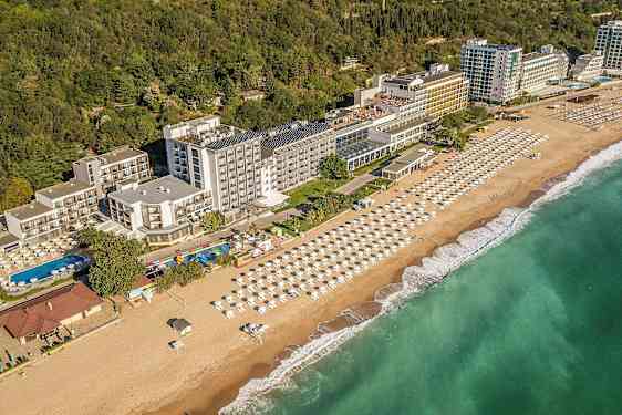 Udsigt over hotel ved kysten med strand og parasoller samt grønne områder i baggrunden. (AI-genereret tekst)