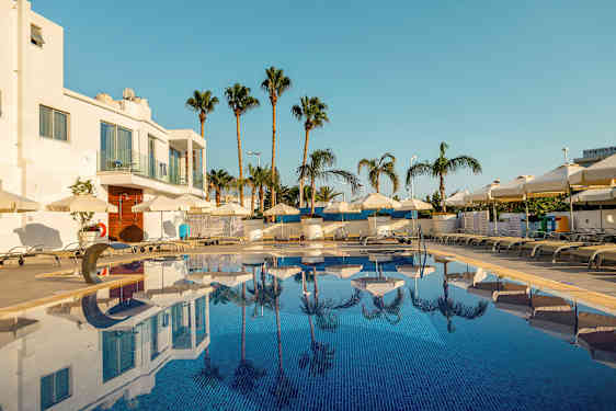 Billede af en udendørs swimmingpool omgivet af liggestole og parasoller, med palmer og en hvid bygning i baggrunden. (AI-genereret tekst)