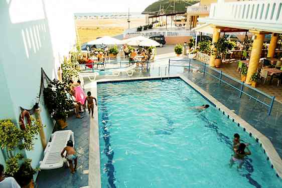 En udendørs pool omgivet af liggestole og borde, med folk der svømmer og hygger sig. (AI-genereret tekst)