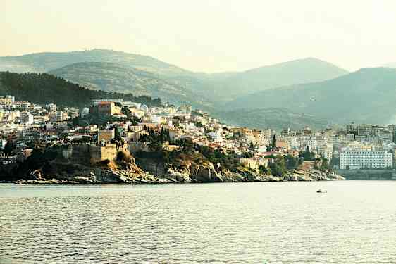 Billede der viser en kystby med bygninger på en bakke nær havet, med både på vandet og grønne bakker i baggrunden. (AI-genereret tekst)