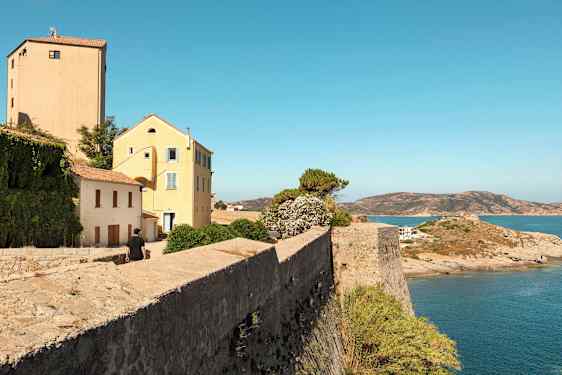 Billede af en stenmur ved vandet med huse og vegetation i baggrunden, samt en mand på vej mod bygningerne. (AI-genereret tekst)