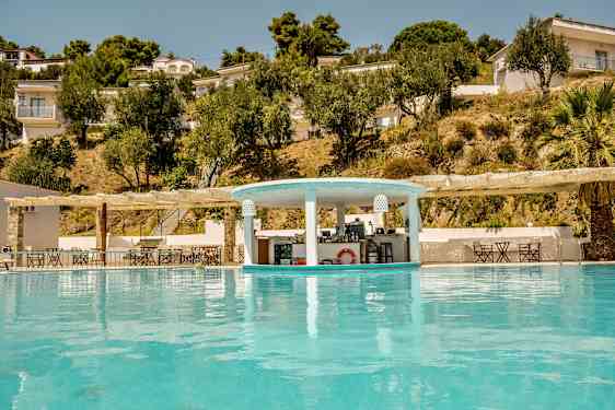 En pool med en poolbar og omkringliggende bygninger og træer på skråningen. (AI-genereret tekst)