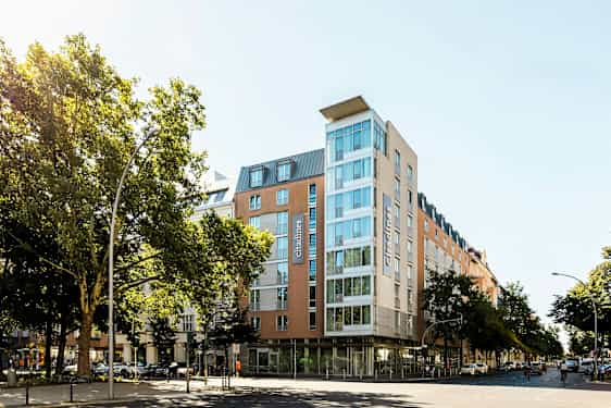 Et moderne hotel med glasfacade omgivet af træer og bymiljø. (AI-genereret tekst)