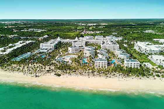 Luftfoto af et hotelkompleks ved en strand, inklusive poolområde, omgivet af grønne omgivelser. (AI-genereret tekst)
