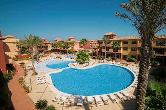 Hotelområde med pool og liggestole, omgivet af palmer og bygninger i varme farver. (AI-genereret tekst)