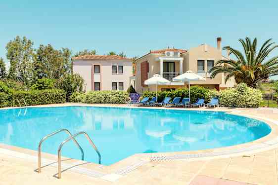 En pool omgivet af liggestole og parasoller, med bygninger og grønt i baggrunden. (AI-genereret tekst)