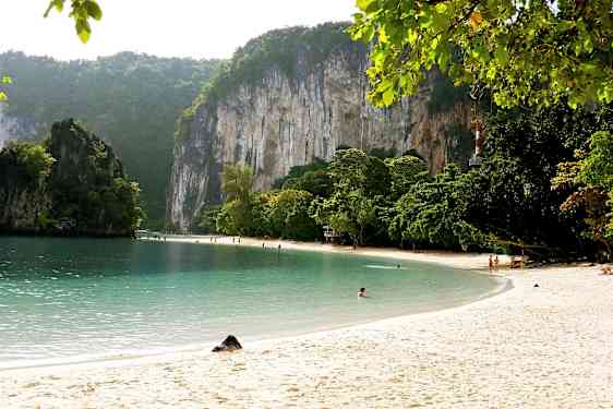 Sandstrand med klart vand, omgivet af grønne træer og høje kalkstensklipper. (AI-genereret tekst)