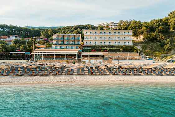 En sandstrand ved havet med liggestole og parasoller. I baggrunden ses hotelbygninger og beplantning. (AI-genereret tekst)