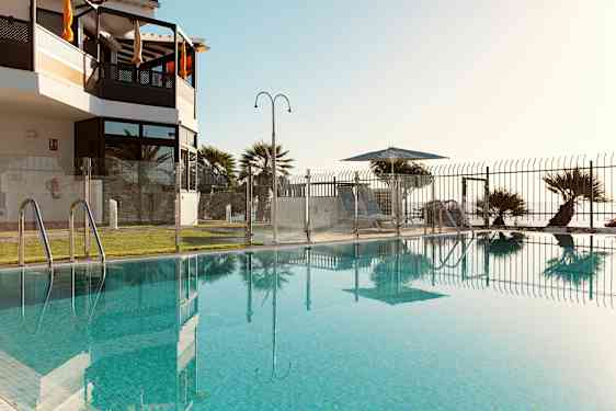 En pool omgivet af et glashegn med udsigt over en græsplæne og liggestole under parasoller. (AI-genereret tekst)