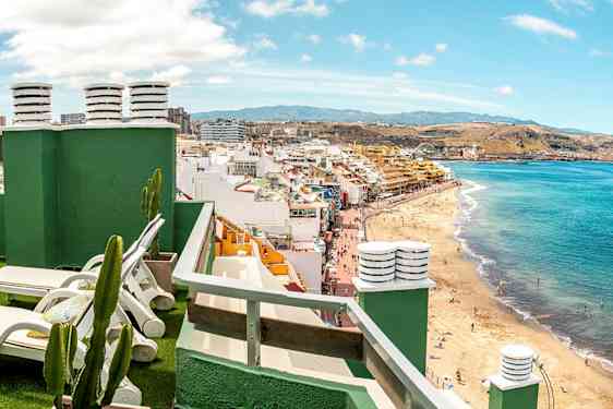 Udsigt fra en tagterrasse med liggestole mod en strand og bymiljø. (AI-genereret tekst)