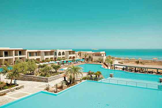 Et ferieresort med poolområde omgivet af tropiske planter og bygninger samt udsigt over havet. (AI-genereret tekst)