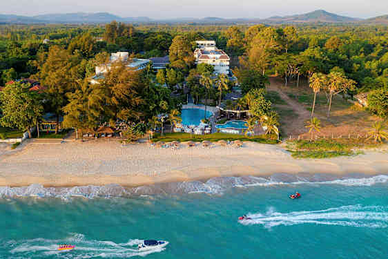 En strand med parasoller og stole samt et hotelkompleks og swimmingpools i baggrunden omgivet af beplantning. (AI-genereret tekst)