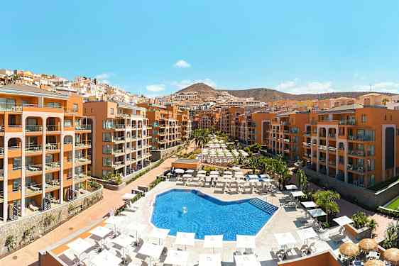 Et hotelområde med en udendørs pool omgivet af liggestole og parasoller samt bygninger i terrakottafarvet. (AI-genereret tekst)