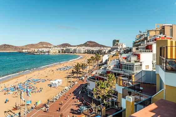 En sandstrand med parasoller nær en by med bygninger og bjerge i baggrunden. (AI-genereret tekst)