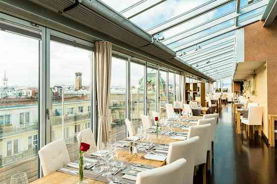 En lys restaurant med glasvægge og glastag, bordene er dækket med hvide stole og en rose på hvert bord. (AI-genereret tekst)