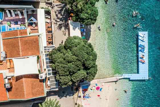 Luftfoto af bygninger med røde tage, en pool, en strand og turkisblåt hav. (AI-genereret tekst)