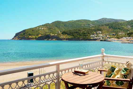 Udsigt fra en balkon mod havet og frodige bakker, med en træstol og et bord. (AI-genereret tekst)