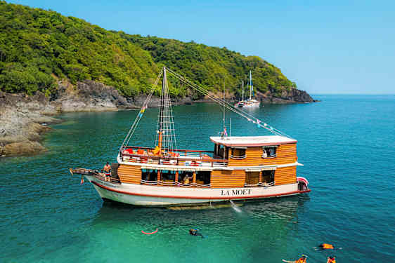 En træbåd med navnet La Moet er fortøjet ved turkisvand nær en skovklædt kyst. (AI-genereret tekst)