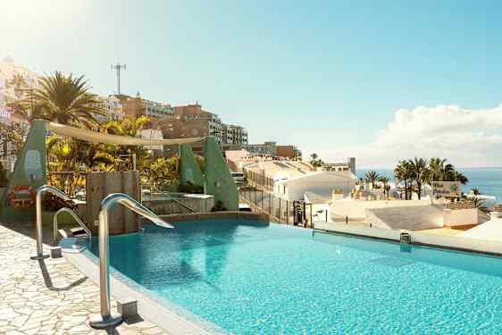 En infinity-pool med udsigt over hustage, palmer og havet i et solrigt landskab. (AI-genereret tekst)