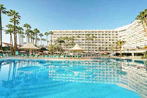 Hotelbygning med stor pool og liggestole i forgrunden, omgivet af palmer. (AI-genereret tekst)