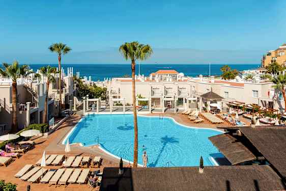 Udsigt over et hotelområde med pool og liggestole, havet synligt i baggrunden. (AI-genereret tekst)