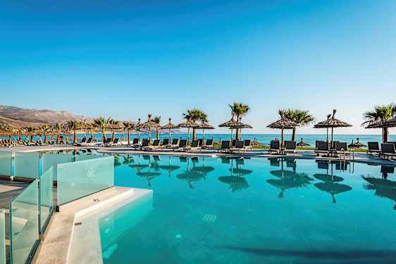 En pool foran stranden med liggestole og parasoller, bjerge i baggrunden. (AI-genereret tekst)