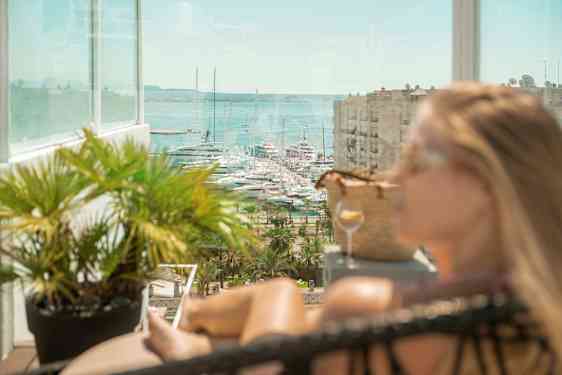 Udsigt mod både i en marina, med en glasaltan og frodige planter i forgrunden. (AI-genereret tekst)