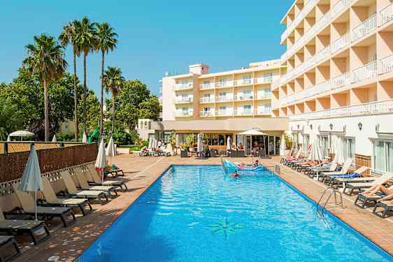 En hotelbygning ses ved siden af en udendørs pool med liggestole og parasoller omkring. (AI-genereret tekst)