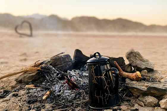 En kaffekande står ved et udbrændt bål i et ørkenlandskab. (AI-genereret tekst)