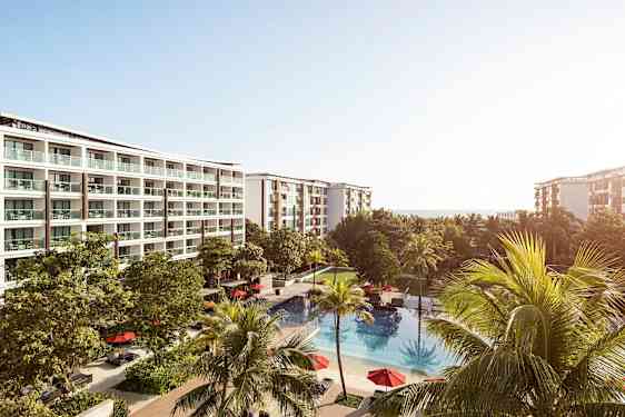 Et hotelkompleks med flere etager og altaner, omgivet af grønne træer. Foran bygningerne ligger en udendørs pool med røde parasoller. (AI-genereret tekst)