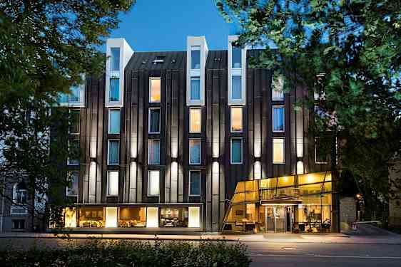 Moderne hotels facade med glas og oplyste vinduer omgivet af træer. (AI-genereret tekst)