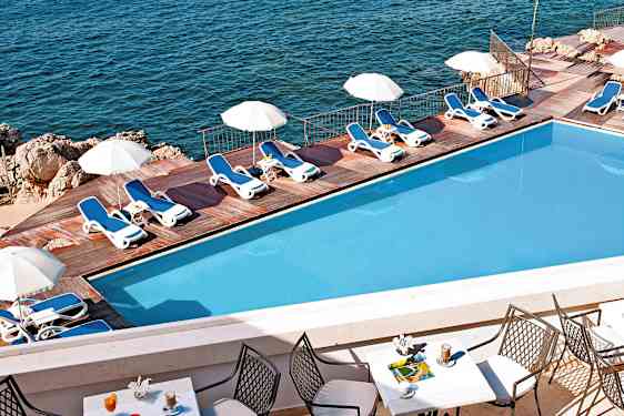 En pool med solsenge og parasoller ved en træterrasse nær havet. Forgrunden viser et bord med stole og drikkevarer. (AI-genereret tekst)