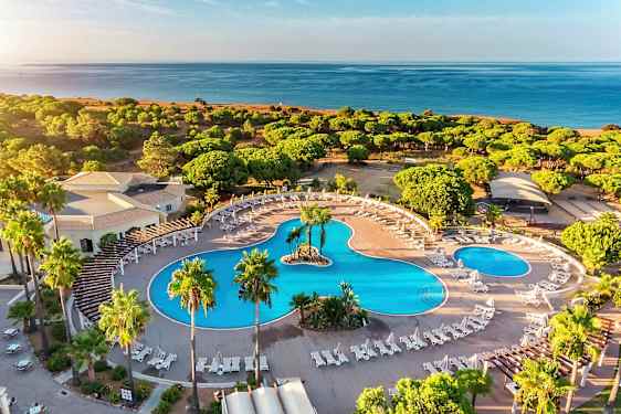 Luftfoto af et hotelområde med store swimmingpools, liggestole og omgivelser med grønne områder nær stranden. (AI-genereret tekst)
