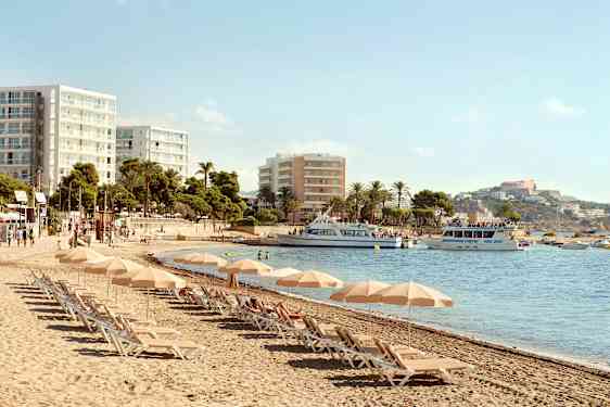 Strand med liggestole og parasoller, hotelbygninger i baggrunden og både på vandet. (AI-genereret tekst)