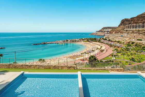 Billede af en strand med parasoller, turkis vand og en pool i forgrunden. (AI-genereret tekst)