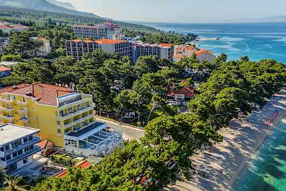 Luftfoto af en strand med liggestole og parasoller ved siden af hotelbygninger og grønne områder. (AI-genereret tekst)