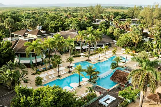 Luftfoto af et hotelområde med en stor pool omgivet af liggestole og parasoller samt frodige træer og vegetation. (AI-genereret tekst)