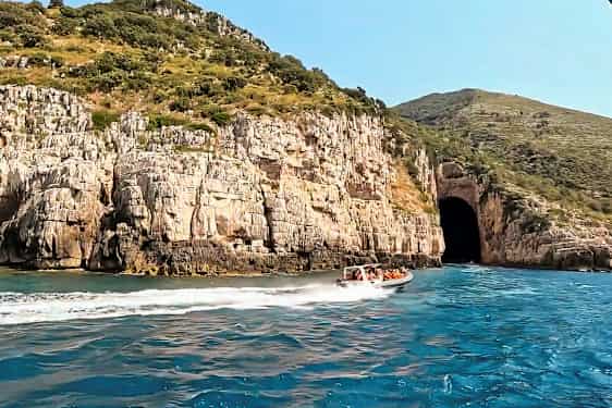 En motorbåd med mennesker sejler på blåt vand tæt på en klippe med en stor grotteåbning. (AI-genereret tekst)