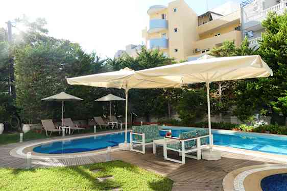 Udendørs pool med liggestole og parasoller, omgivet af træer og bygninger. (AI-genereret tekst)