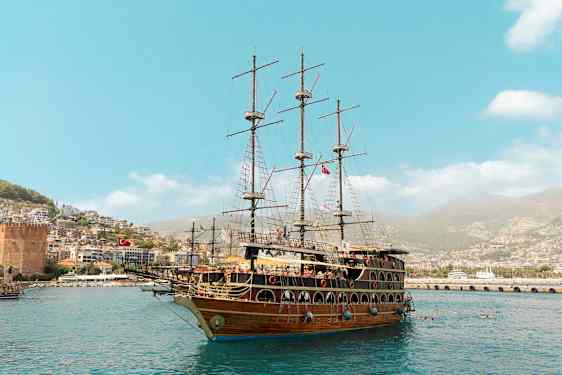 Et stort sejlskib ligger i havnen med en by og grønne bakker i baggrunden. (AI-genereret tekst)