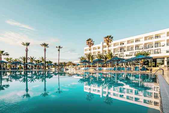 Udvendig af et hotel med en stor pool foran og omgivende palmer. (AI-genereret tekst)