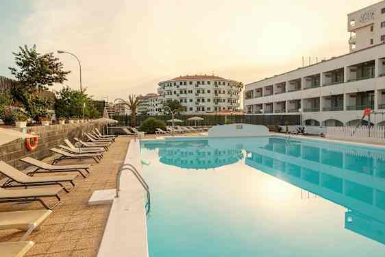 Udsigt over en pool med solsenge ved siden af og omkringliggende bygninger. (AI-genereret tekst)