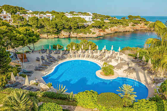 En udendørs pool med liggestole og parasoller, omgivet af grønt og med udsigt til havet. (AI-genereret tekst)