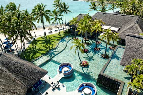 Flere huse med stråtag ved en pool med palmer og sandstrand i baggrunden. (AI-genereret tekst)