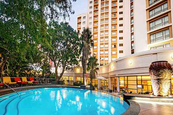 En oplyst udendørs pool omgivet af grønne planter og en bygning i baggrunden. (AI-genereret tekst)