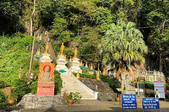 Billede af en trappe, der fører op til flere gyldne buddhistiske statuer og strukturer omgivet af greenery. (AI-genereret tekst)