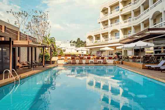 En udendørs swimmingpool med liggestole og parasoller ved siden af og et fler-etagers hotel i baggrunden. (AI-genereret tekst)