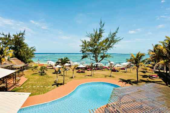 En pool med turkis vand foran en græsplæne med træer, liggestole og parasoller langs en strand. (AI-genereret tekst)