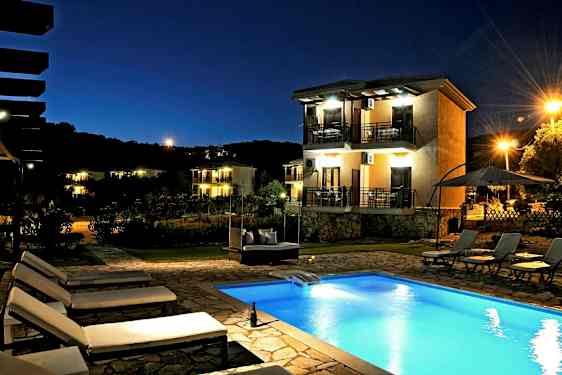 Et aftenbillede af en udendørs pool og flere liggestole. I baggrunden ses et hus med altaner og omgivende belysning. (AI-genereret tekst)