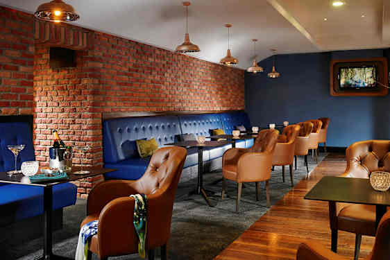 En restaurant eller lounge med blåpolstrede bænke og brune læderstole. Væggene er af røde mursten, og der er små borde med dekorative lys. (AI-genereret tekst)